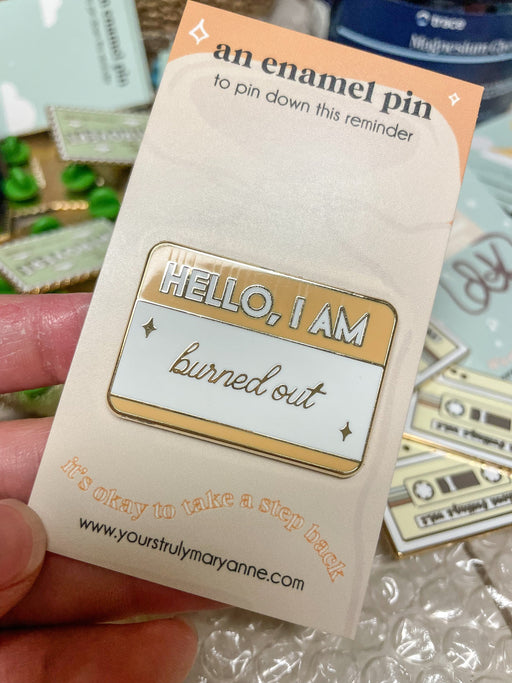 Name-tag enamel pin reading HELLO, I AM burned out, with a warm yellow header, pale background, and star accents.
