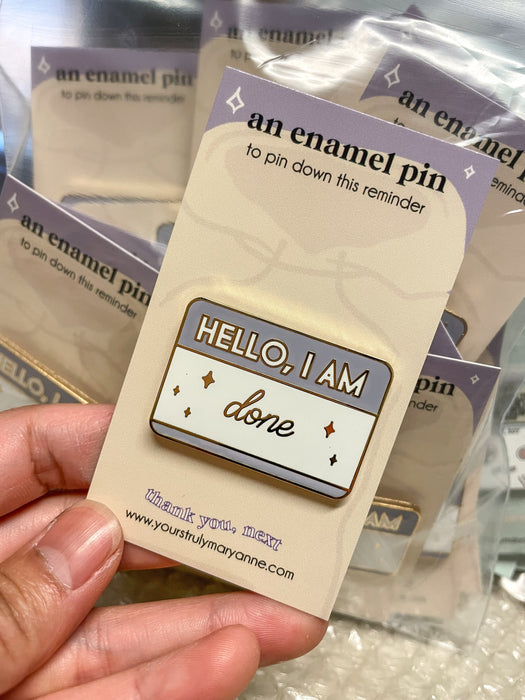 Lavender-and-cream name-tag enamel pin reading HELLO, I AM done, with metallic outlines and star accents.