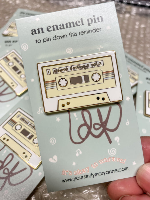 Cream-and-white cassette tape enamel pin reading Mixed feelings vol. 1 on a Yours Truly, Mary Anne backing card.